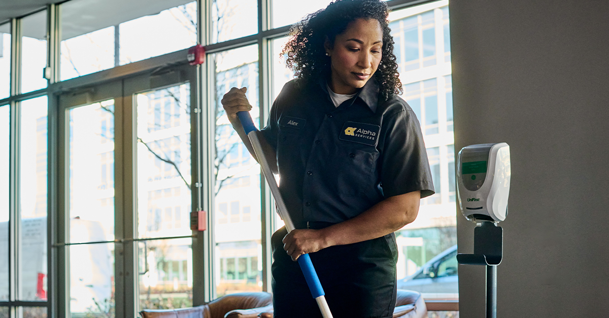 Woman in UniFirst grey polo shirt, looking down and using mop to clean floor