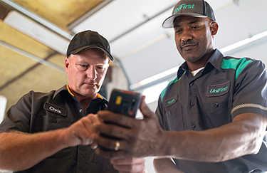 A UniFirst RSR standing with a customer looking at his handheld and working on UniFirst's uniform rental service