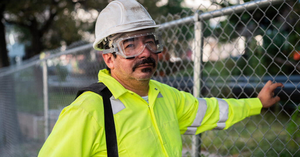 A man standing next to a chain link fence wearing UniFirst high visibility garments and PPE