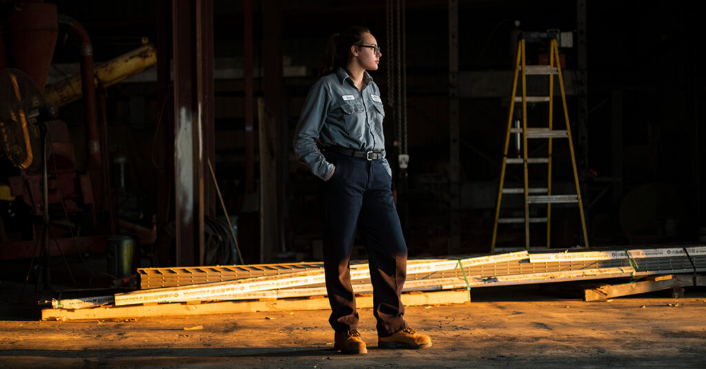 A woman stands in a warehouse with construction equipment in the background wearing a UniFirst industrial uniform.