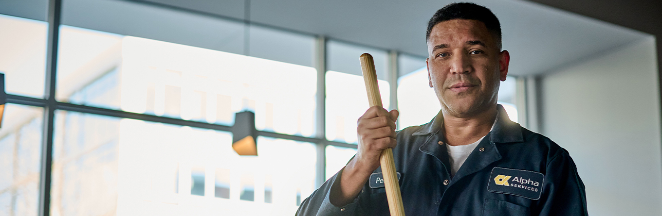 A man standing in the lobby of a building smiling and holding a mop handle while wearing a uniform coverall