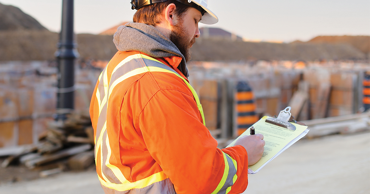 A construction worker wearing high visibility clothing writing on a clipboard at a job site.